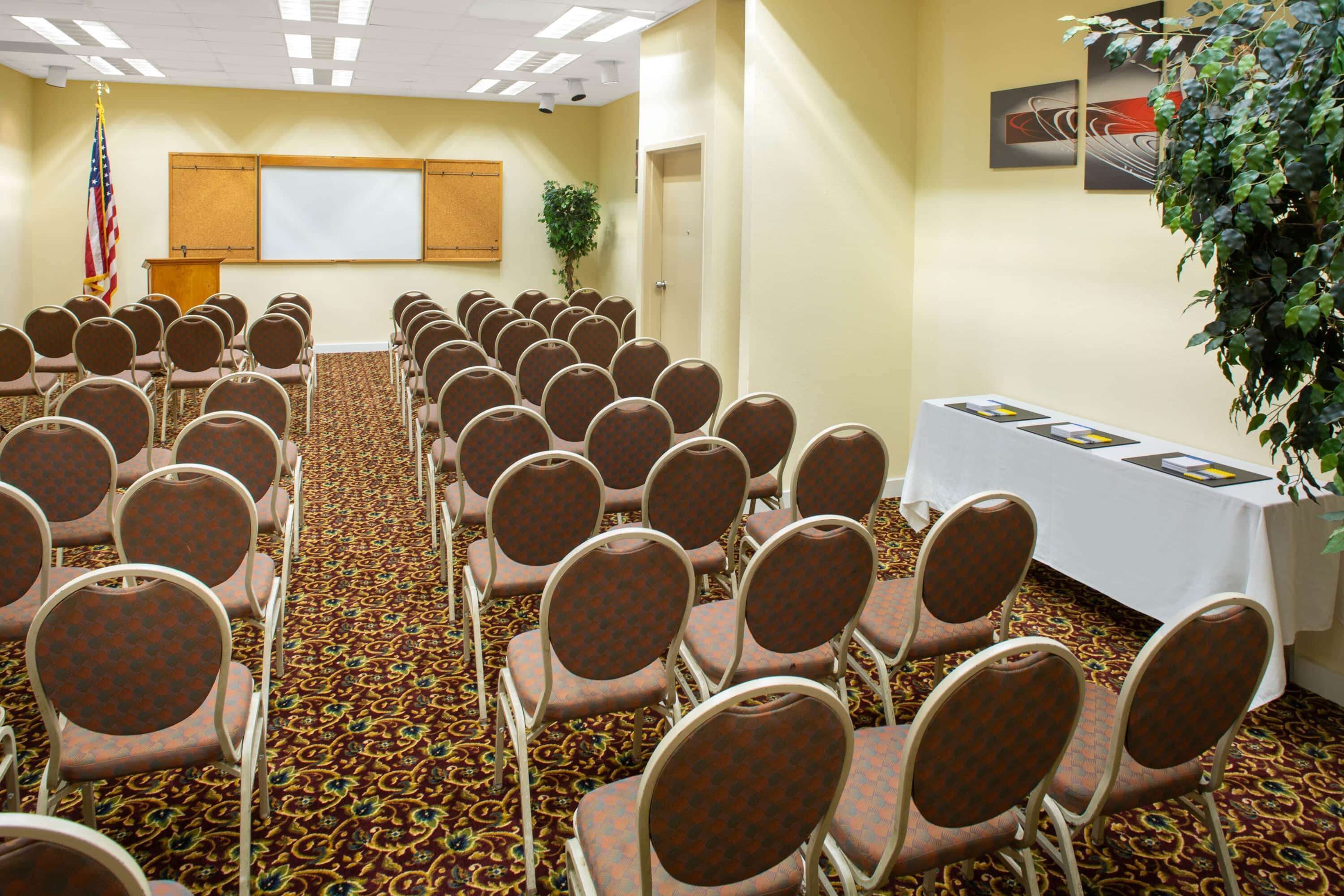 A conference room with rows of chairs, a podium, a projector screen, and a table with brochures.