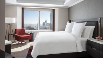 A modern hotel room with a large bed, a red armchair, a desk, and a view of the cityscape through a large window.