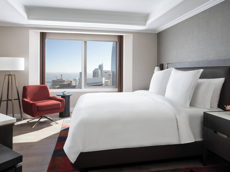 A modern hotel room with a large bed, a red armchair, a desk, and a view of the cityscape through a large window.