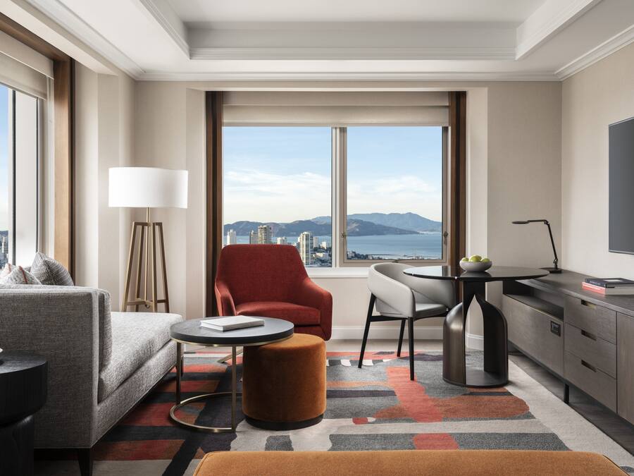 A modern hotel room with a sofa, armchair, dining table, and a TV.