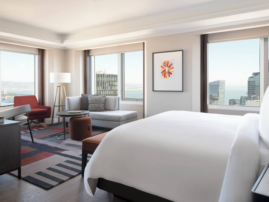 Una habitación de hotel moderna con una cama grande, un escritorio, un sillón rojo, un sofá gris y vistas a la ciudad.