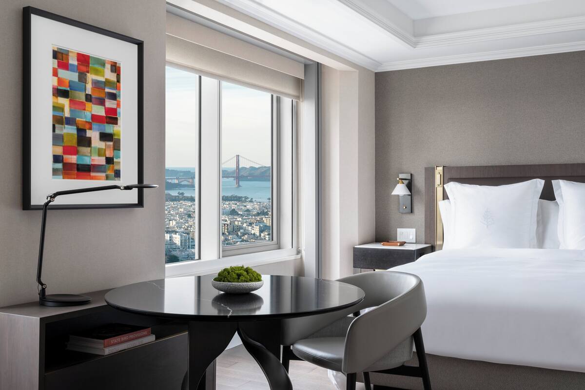 Un cuarto de hotel con una cama grande, una mesa redonda con un recipiente de follaje, un escritorio con libros, una obra de arte enmarcada y vistas a un puente y al paisaje urbano.