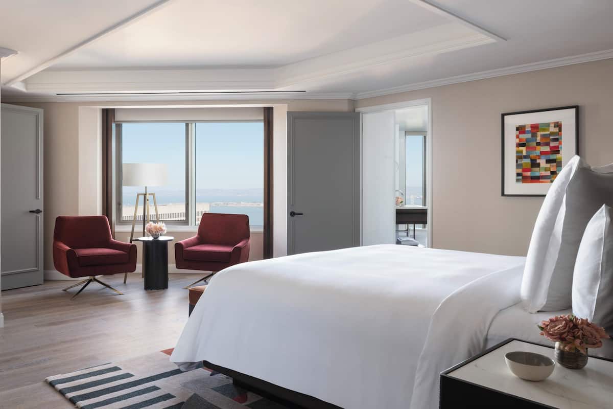 Habitación de hotel moderna con una cama grande, dos sillones rojos, una mesita con una lámpara y vistas a la ciudad.