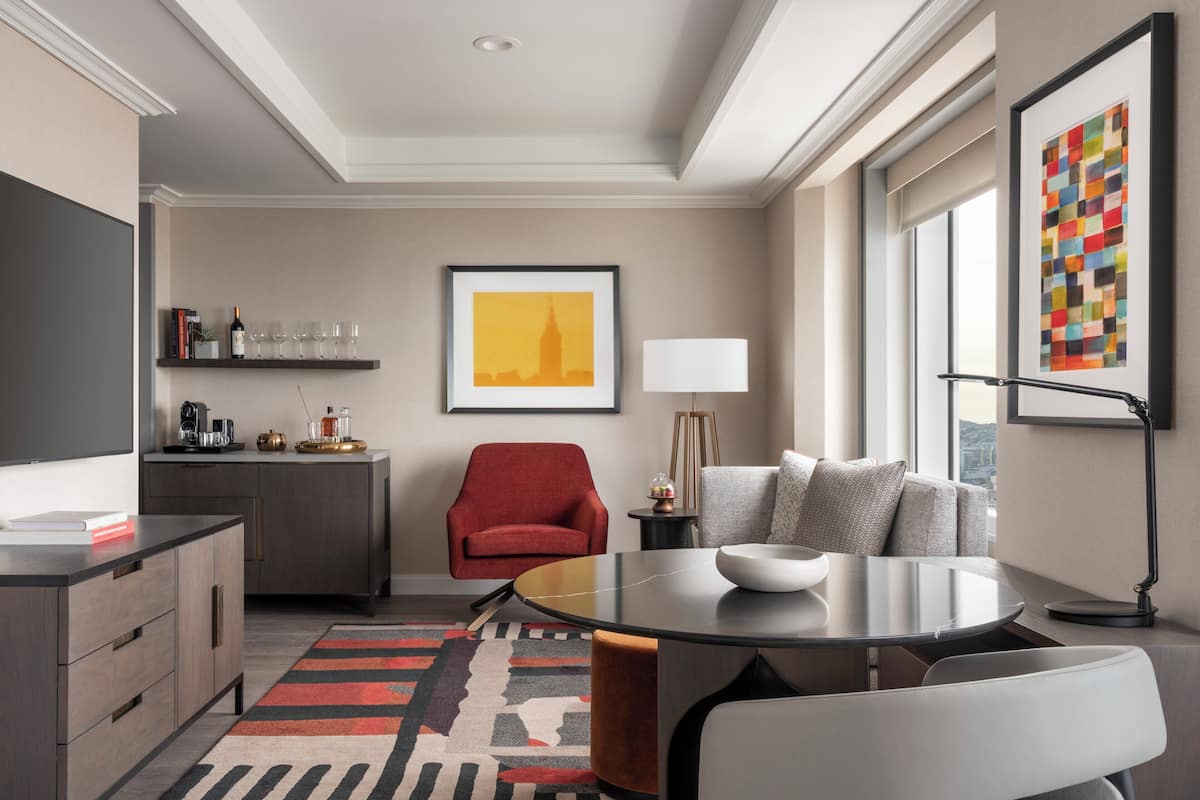 Una habitación de hotel moderna con zona de comedor, un sillón rojo, un sofá, una obra de arte con un motivo urbano y una lámpara.