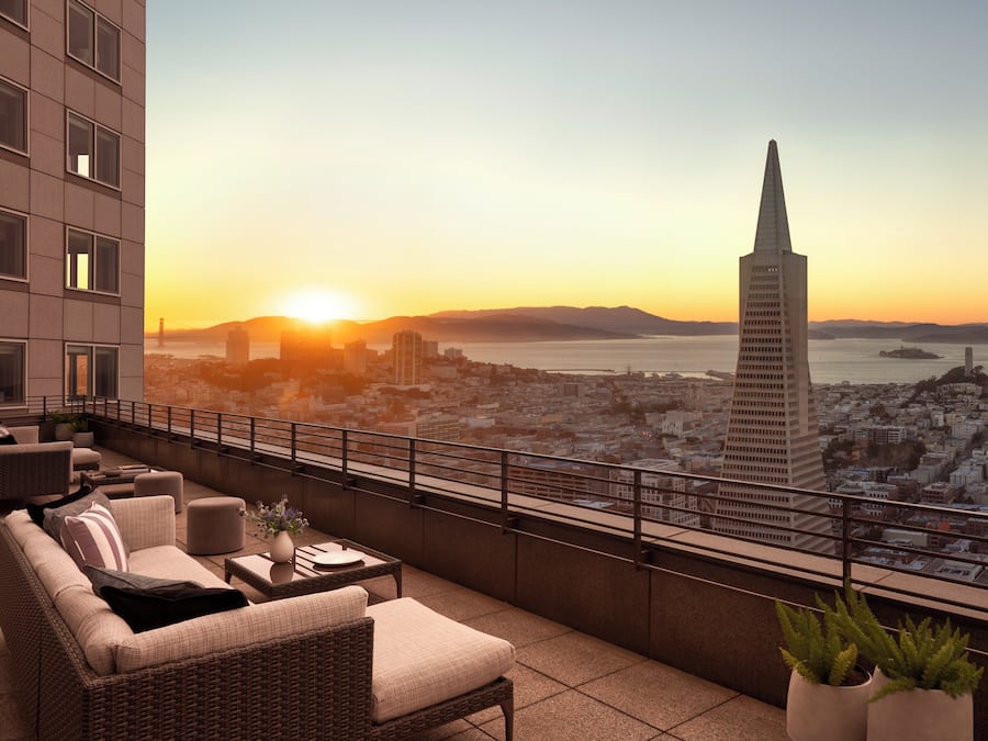 Uma varanda com móveis de vime, plantas em vasos e vista para a cidade ao pôr do sol.