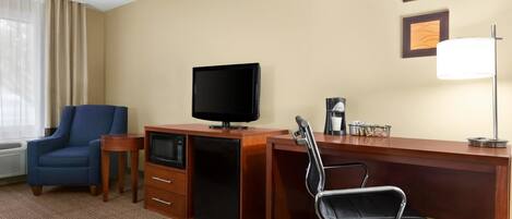 A hotel room with a wooden desk, a television, a blue chair, a desk lamp, and a coffee maker.