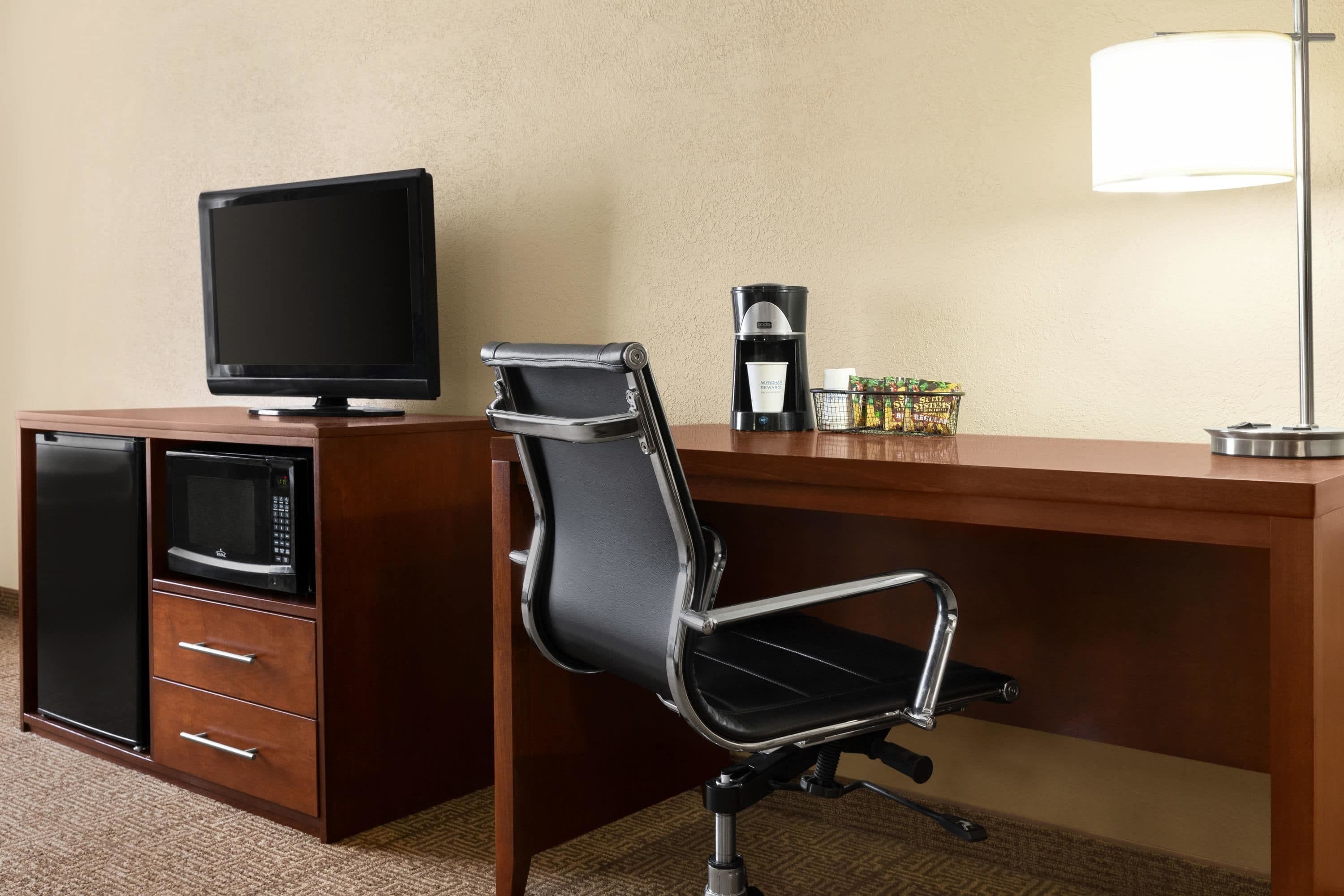 A hotel room with a wooden desk, a black office chair, a television, a coffee maker, and a microwave.