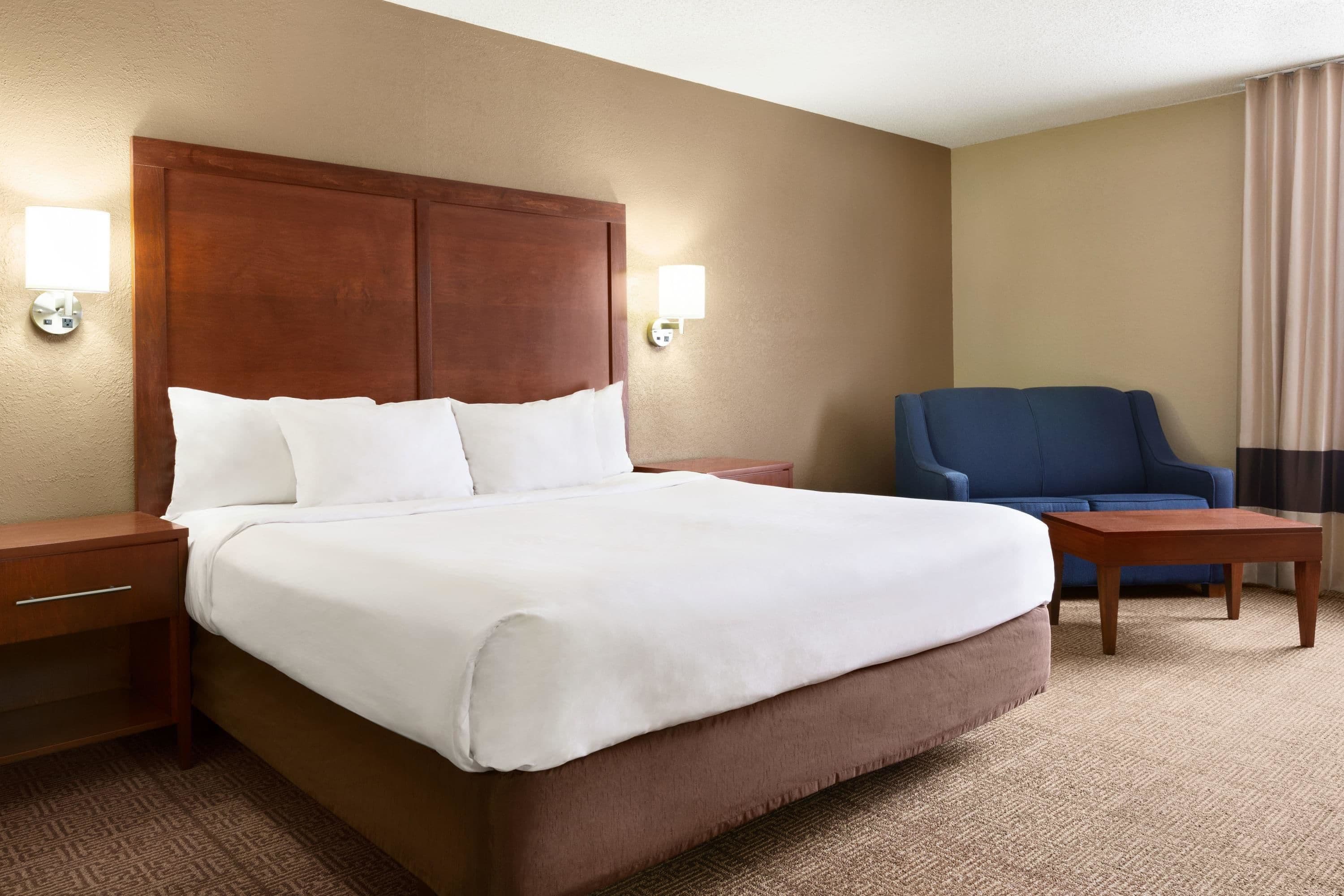 A hotel room with a large bed, two bedside tables, a wooden headboard, a blue armchair, and a small wooden table.