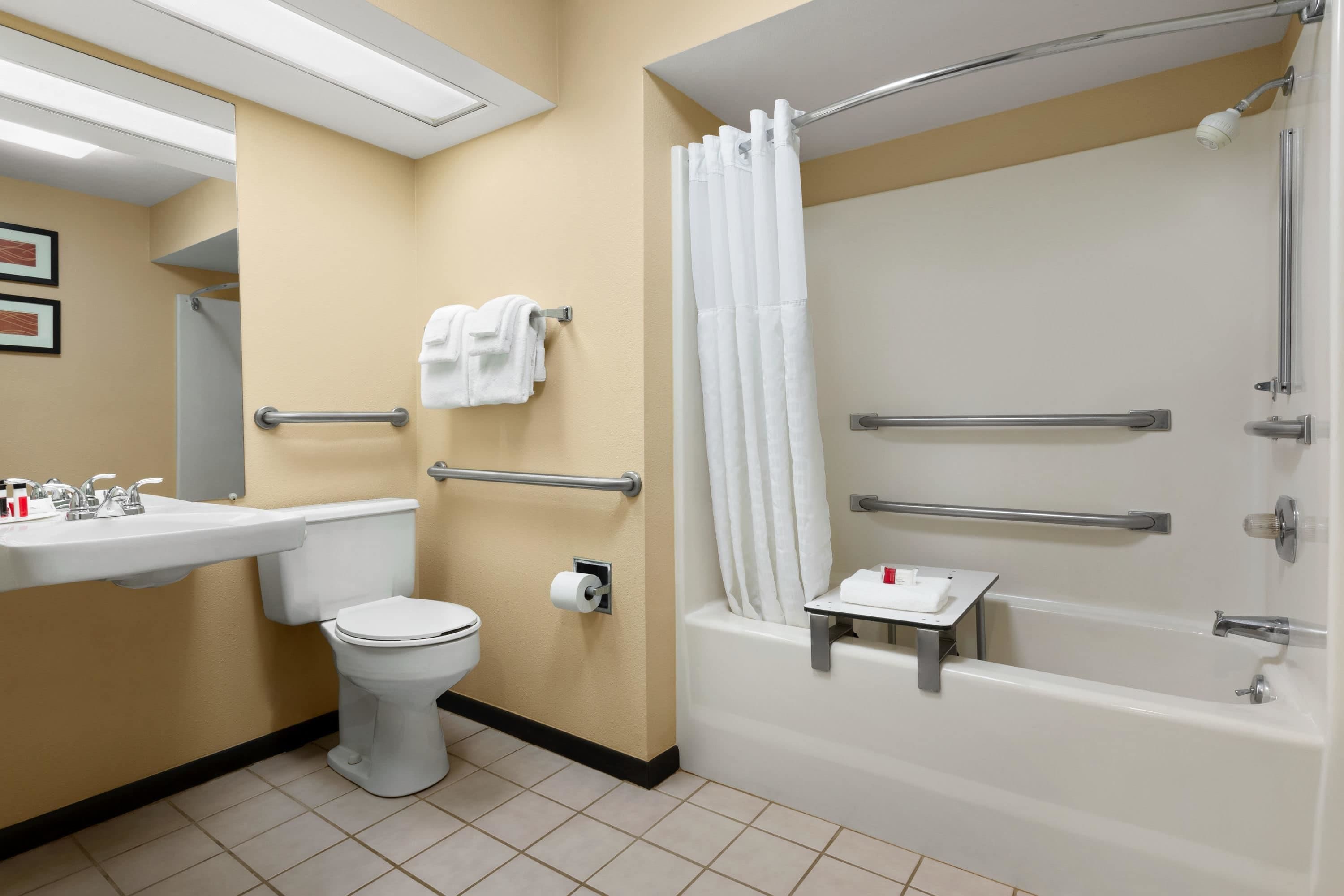 Ein Badezimmer mit Toilette, Waschbecken und Badewanne. Es sind Haltegriffe und eine Duschvorhang vorhanden.