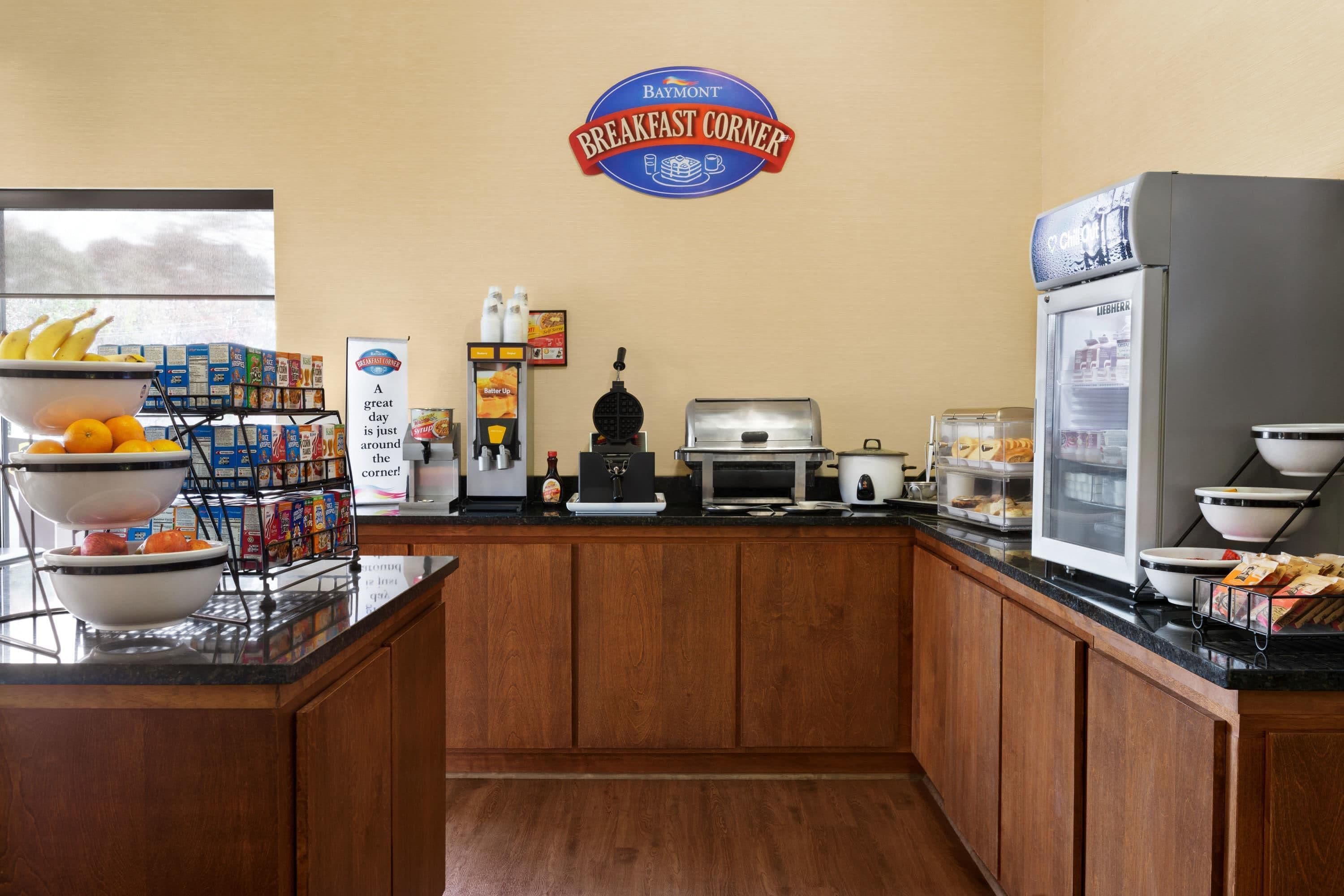 Un área de desayuno con un letrero de Baymont Breakfast Corner, una máquina de café, un refrigerador y varios alimentos para el desayuno en la encimera.