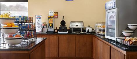 Un área de desayuno con un letrero de Baymont Breakfast Corner, una máquina de café, un refrigerador y varios alimentos para el desayuno en la encimera.