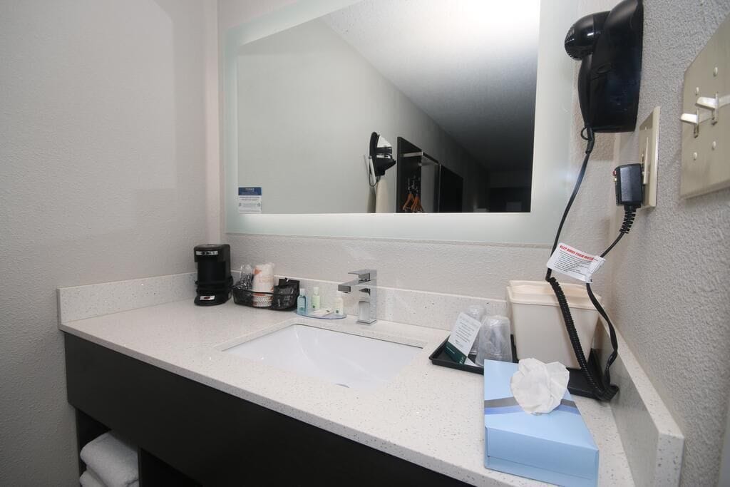 A hotel bathroom with a white sink, a mirror, a hairdryer, and toiletries.
