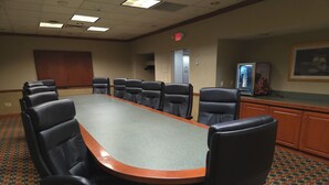 Uma sala de conferências com uma mesa comprida e várias cadeiras, um tapete estampado e um armário com uma televisão.