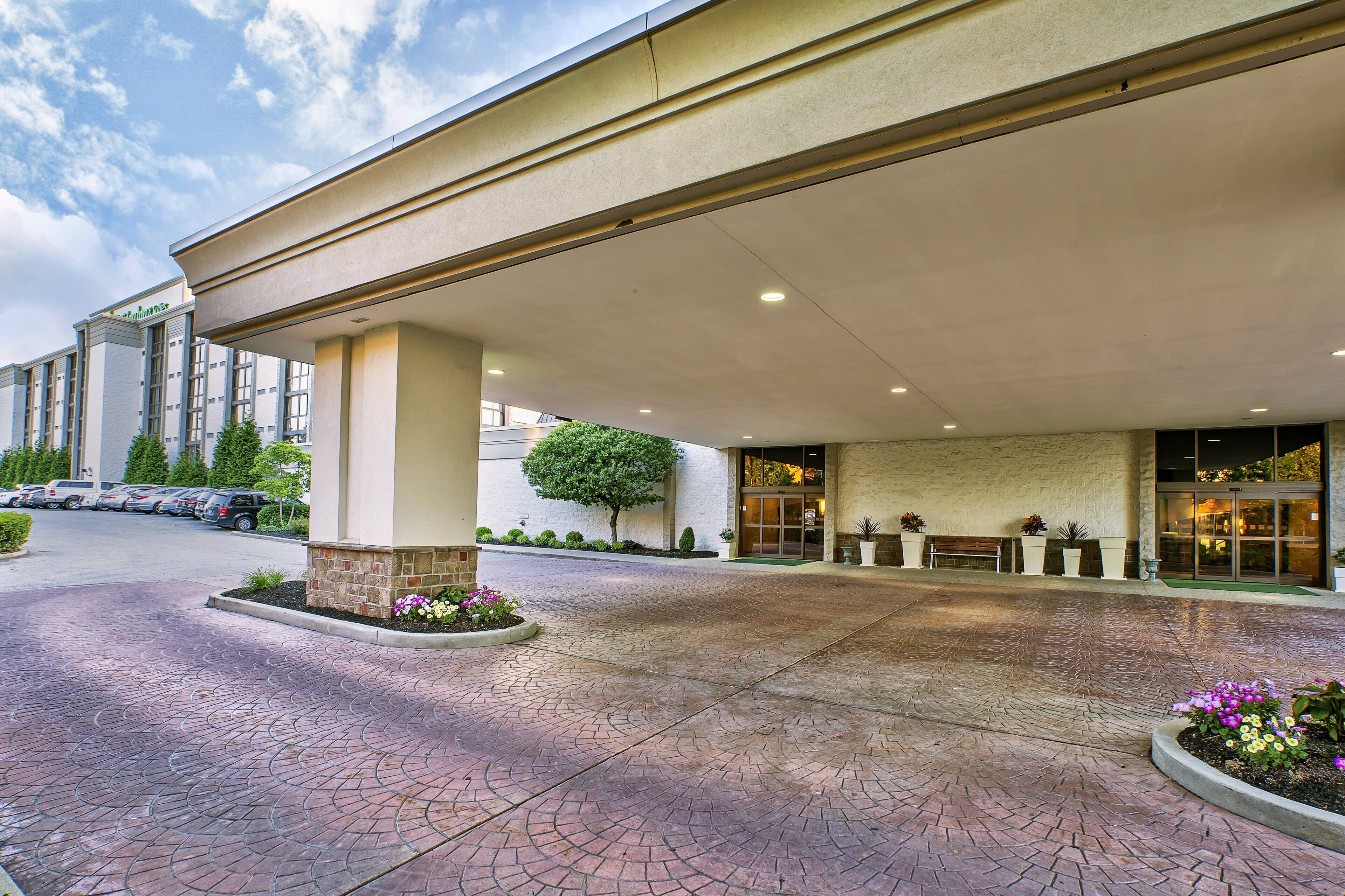 A hotel entrance with a covered walkway, a parking lot, and a landscaped area with flowers.