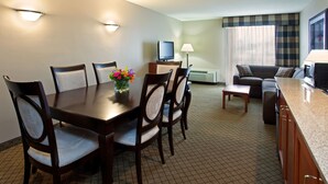 Quarto de hotel com uma mesa de jantar, cadeiras, sofá, televisão, luminária e micro-ondas.