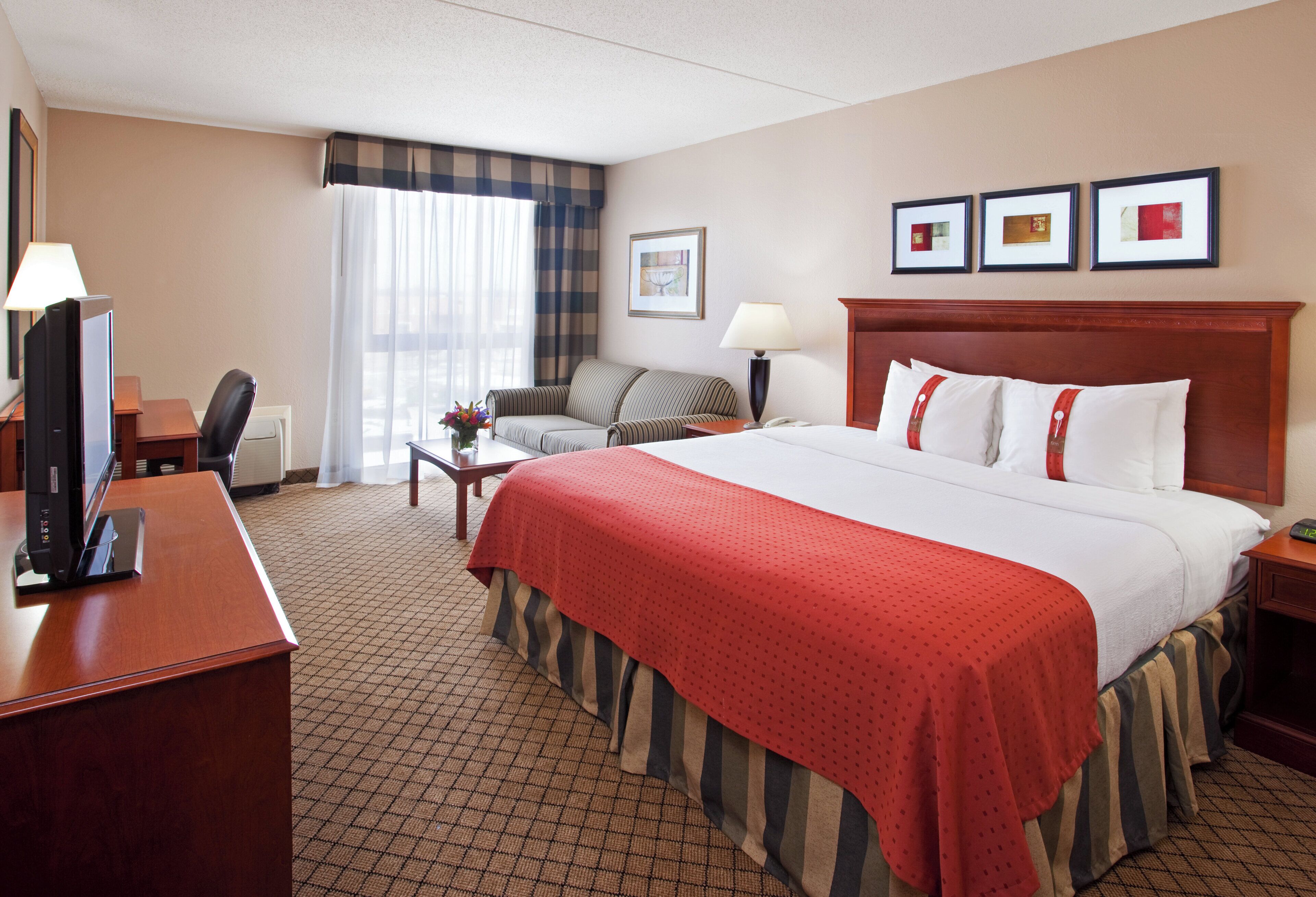 A hotel room with a large bed, a desk with a computer, a chair, a lamp, and a window with curtains.