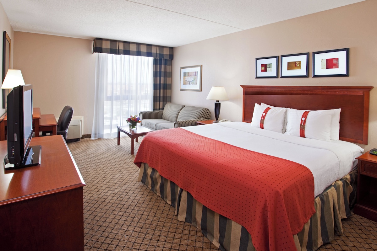A hotel room with a large bed, a desk with a computer, a chair, a lamp, and a window with curtains.