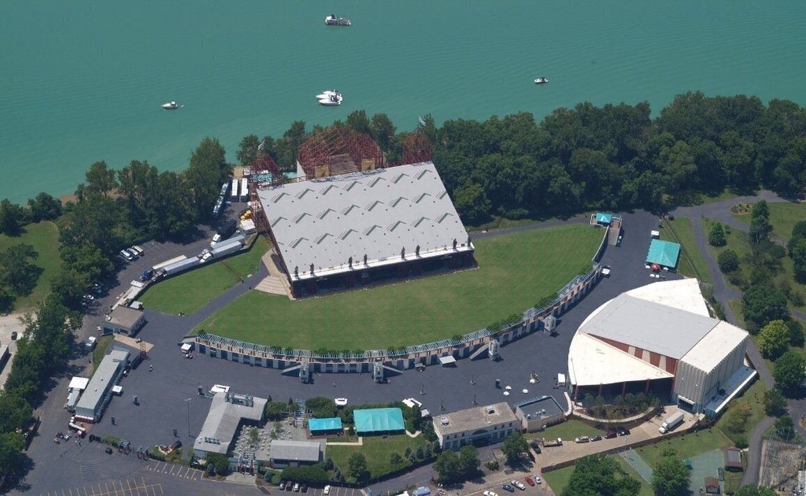 An aerial view of a sports complex with a large stadium, parking areas, and a lake with boats.