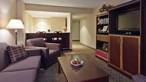 Quarto de hotel com sofá, poltrona, mesa de jantar com maçãs, televisão em um móvel e estante de livros.