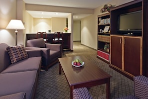 A hotel room with a sofa, armchair, dining table with apples, a TV on a cabinet, and a bookshelf.