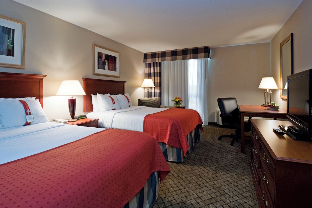 A hotel room with two beds, a desk with a television, a chair, and a lamp.