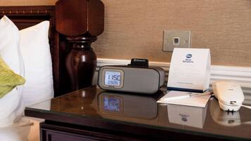 Habitación de hotel con un reloj digital, una mesita de noche con un teléfono, una almohada y un cartel con el nombre del hotel.
