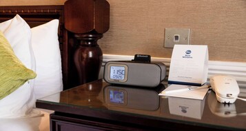 Habitación de hotel con un reloj digital, una mesita de noche con un teléfono, una almohada y un cartel con el nombre del hotel.