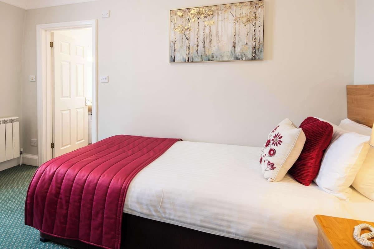 A neatly made bed with a red blanket, white pillows, and a wooden bedside table with a lamp and a telephone.