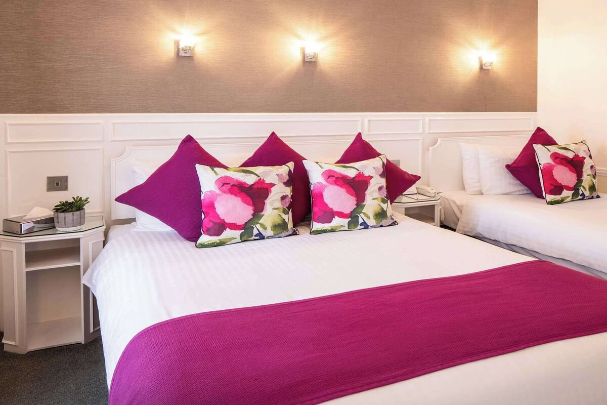 A hotel room with two beds, each with a white headboard and a floral pillow. There are wall-mounted lights and a framed picture on the wall.