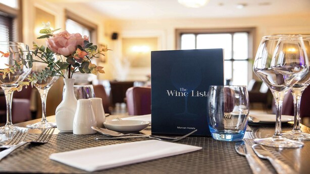 Un tavolo da pranzo in hotel apparecchiato con bicchieri da vino, un menu intitolato "The Wine List", condimenti e un vaso con fiori.