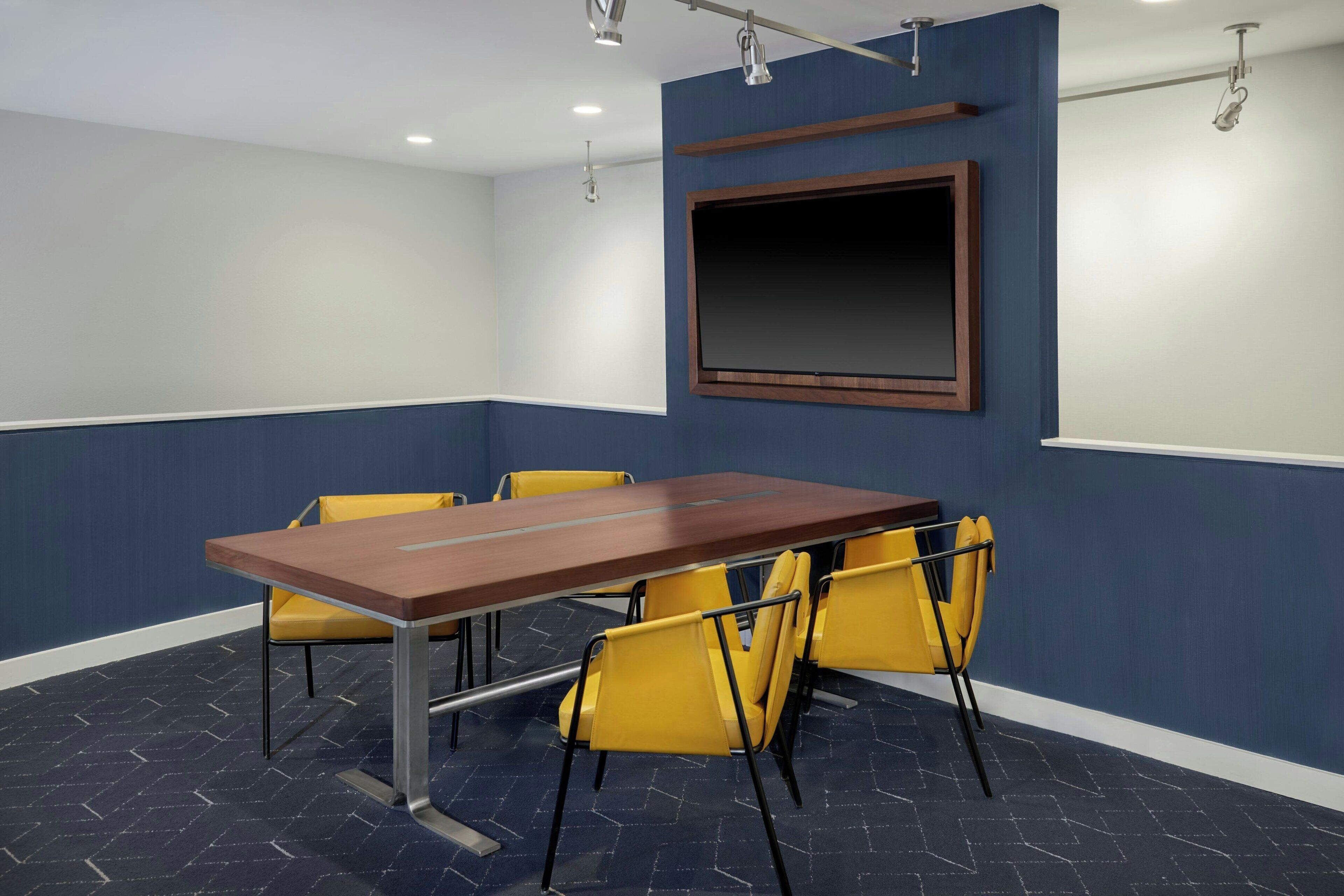 A conference room with a long table, yellow chairs, a large screen, and a blue and white color scheme.