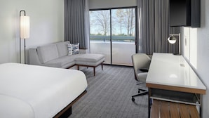 A hotel room with a bed, a sofa, a desk, and a chair. There is a large window with curtains, a flat-screen TV mounted on the wall, and a floor lamp. - Courtyard by Marriott Huntsville University Drive (Huntsville)