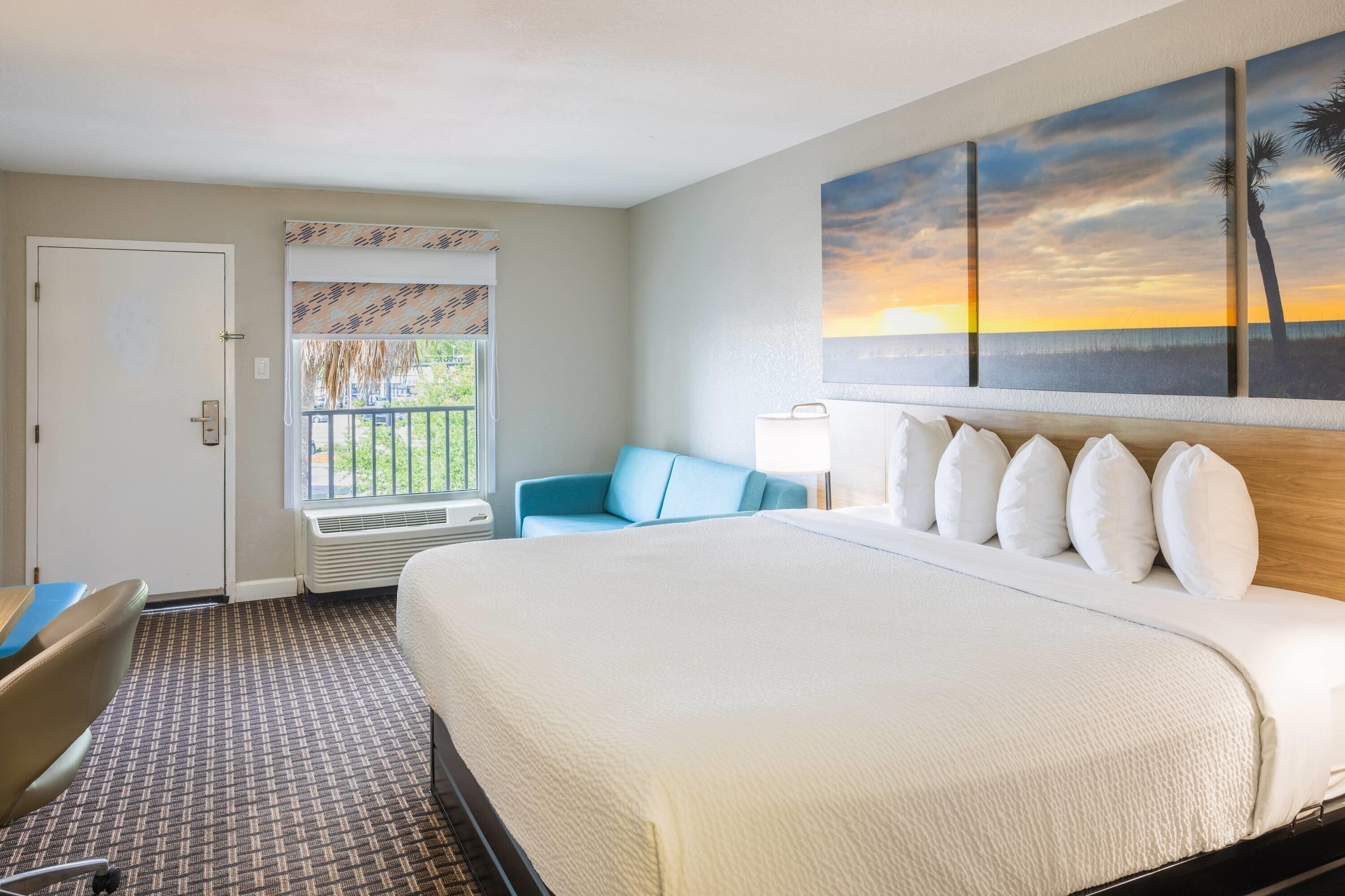Un cuarto de hotel con cama, escritorio, silla, ventana con vista al atardecer y un cuadro en la pared.