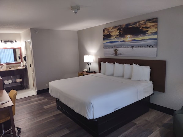 A hotel room with a large bed, a desk with a chair, a nightstand with a lamp, and a painting on the wall.