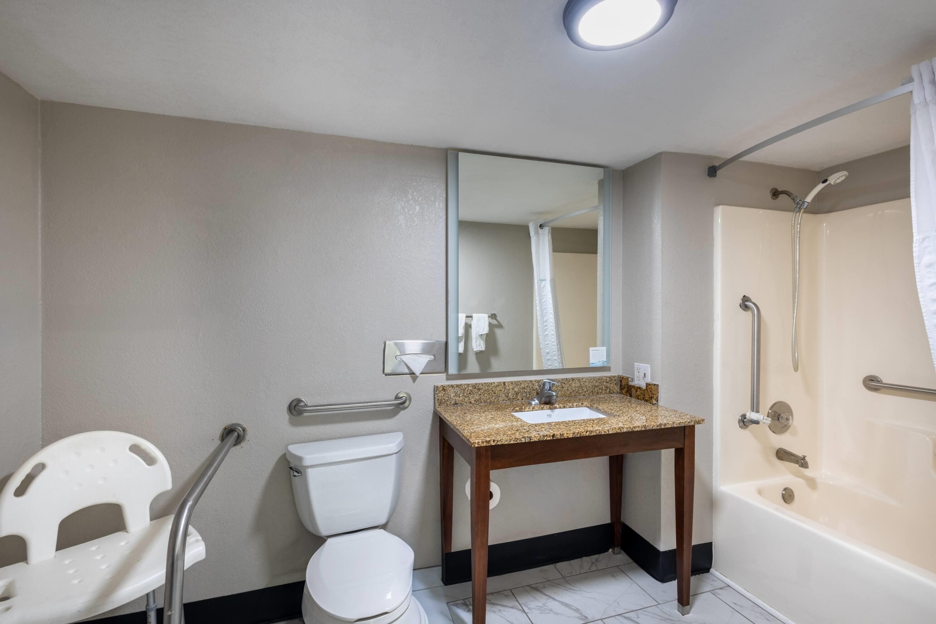 A bathroom with a toilet, sink, mirror, shower, and grab bars.