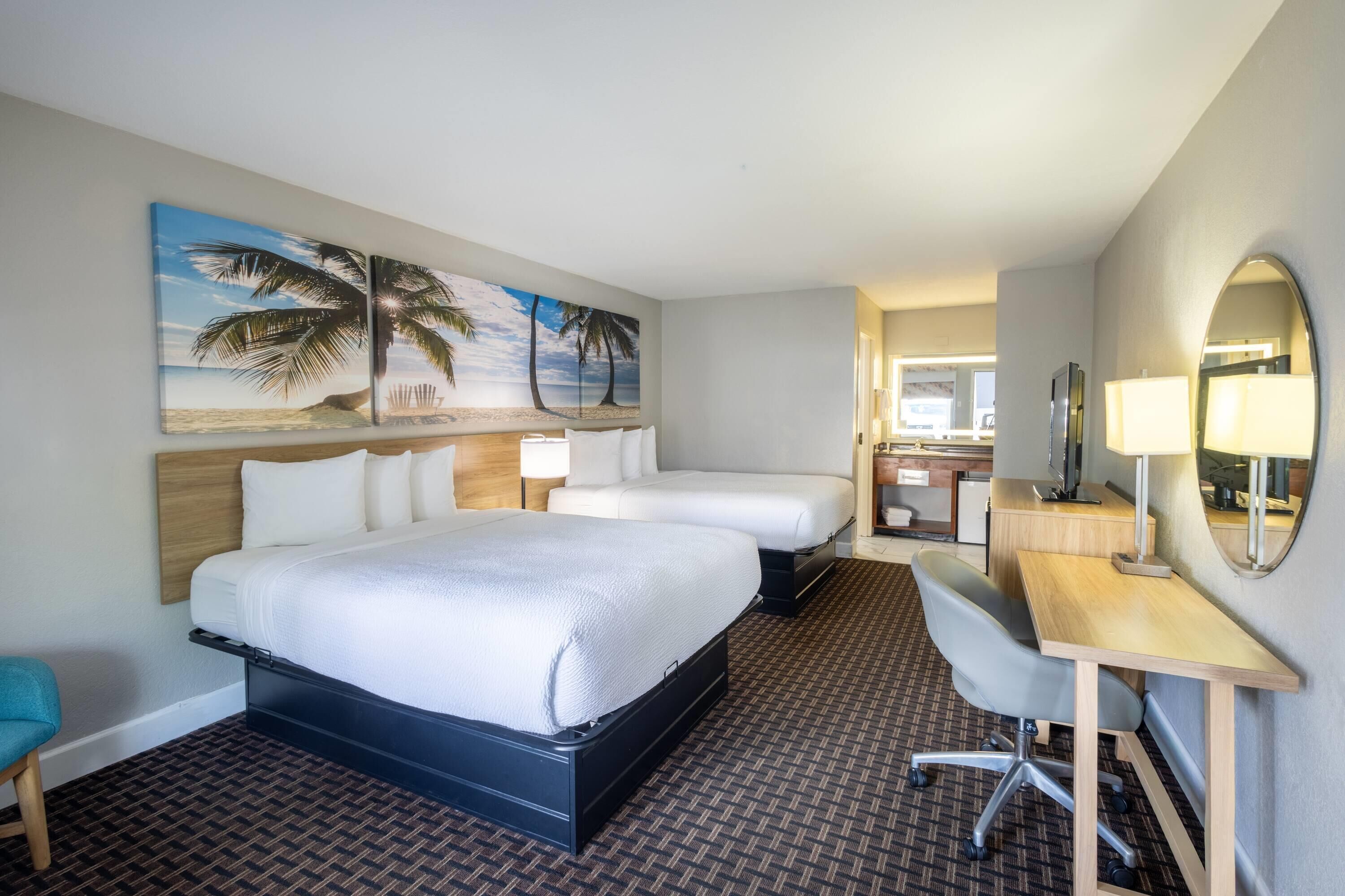 Une chambre d’hôtel comprenant un lit, deux tables de chevet, un bureau, une chaise, un grand miroir et un tableau sur le thème de la plage accroché au mur.