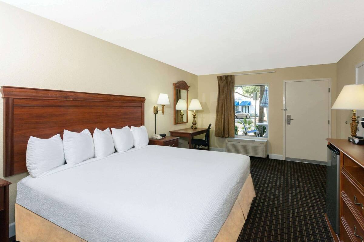 Habitación de hotel con una cama grande, un escritorio con una silla, un espejo, una ventana con cortinas y una puerta.