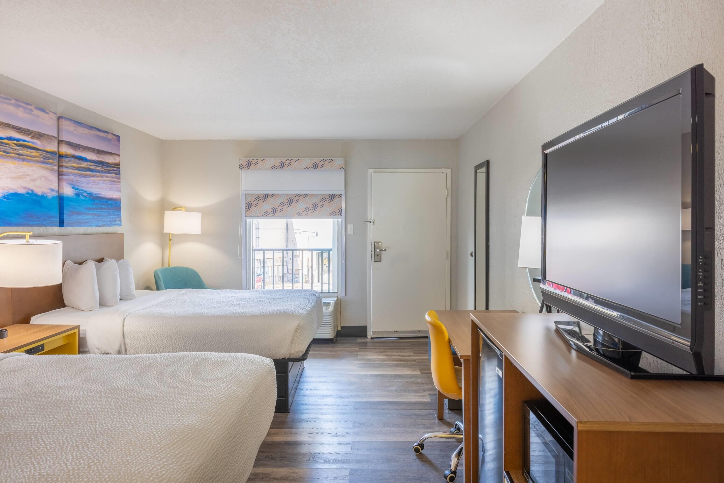 Ein Hotelzimmer mit zwei Betten, einem Fernseher, einem Schreibtisch, einem Stuhl, einer Lampe und einem Fenster mit Jalousien.