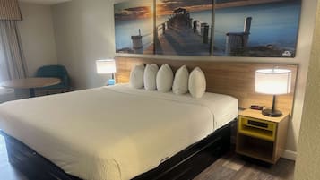 A hotel room with a large bed, a wooden headboard, a nightstand with a lamp, and a painting of a pier on the wall.