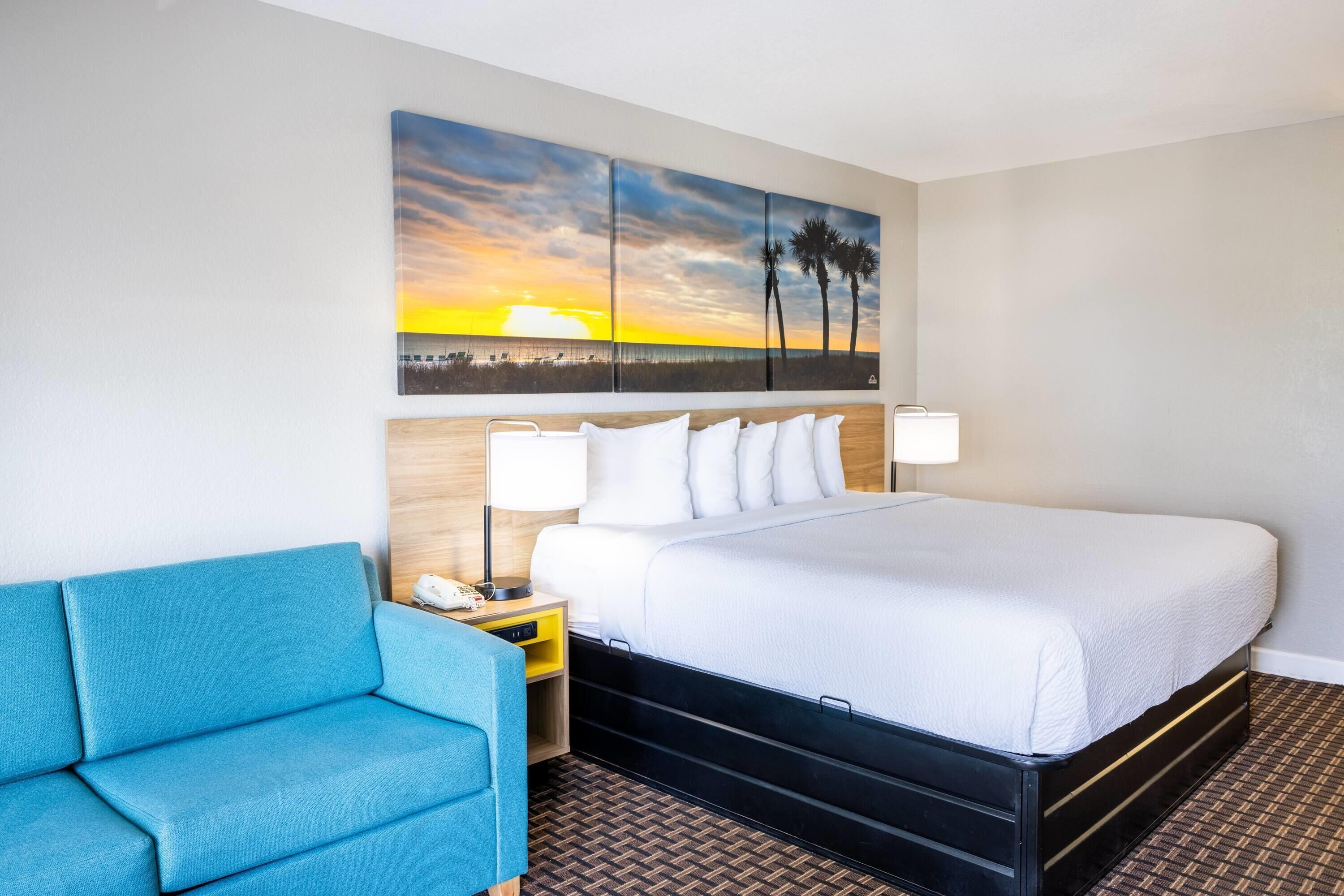 Une chambre d’hôtel avec un lit, un fauteuil bleu, une table de chevet avec une lampe et un mur orné d’un tableau représentant un coucher de soleil.