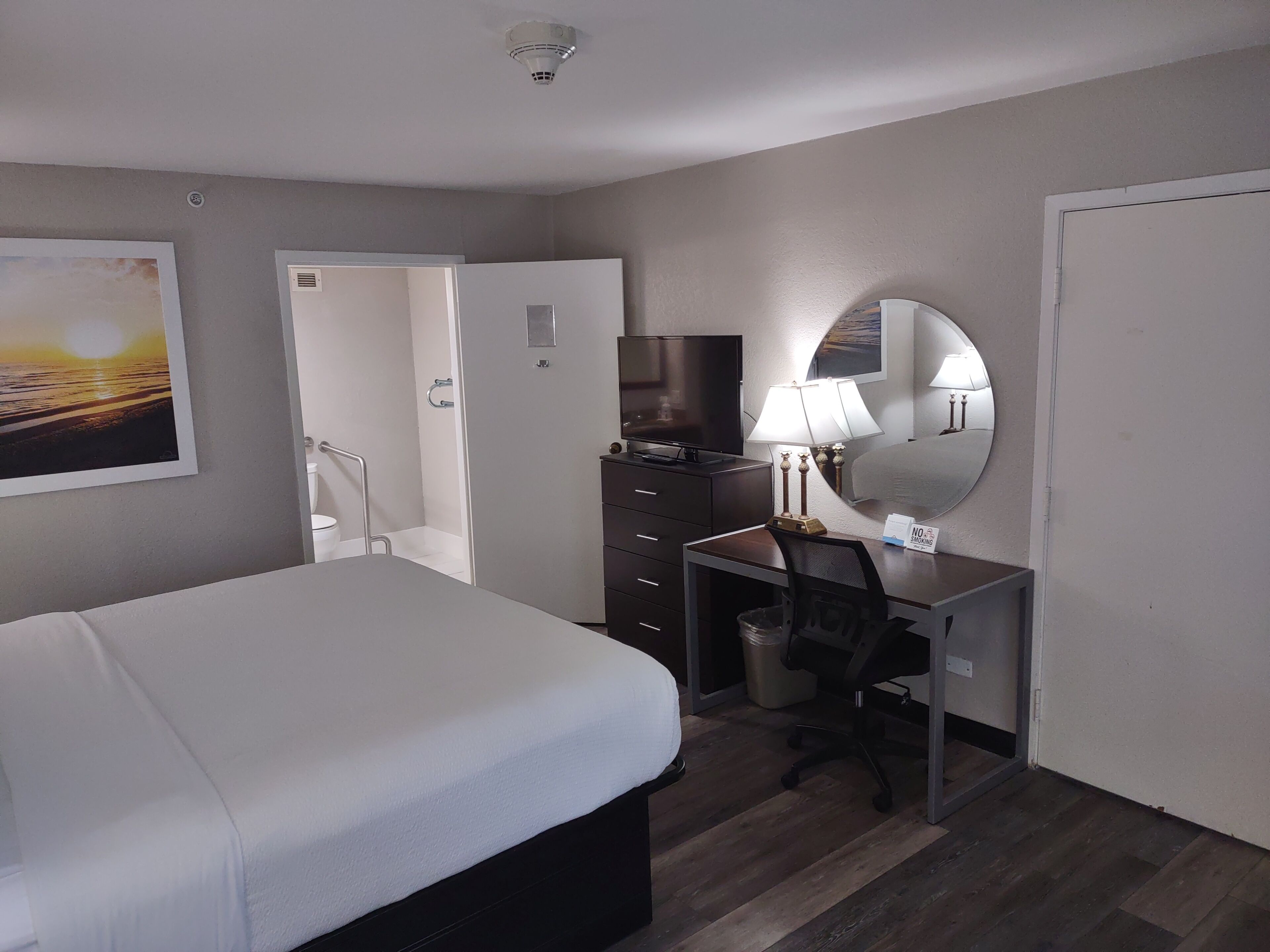 A hotel room with a bed, a desk with a chair, a television, a lamp, and a mirror.