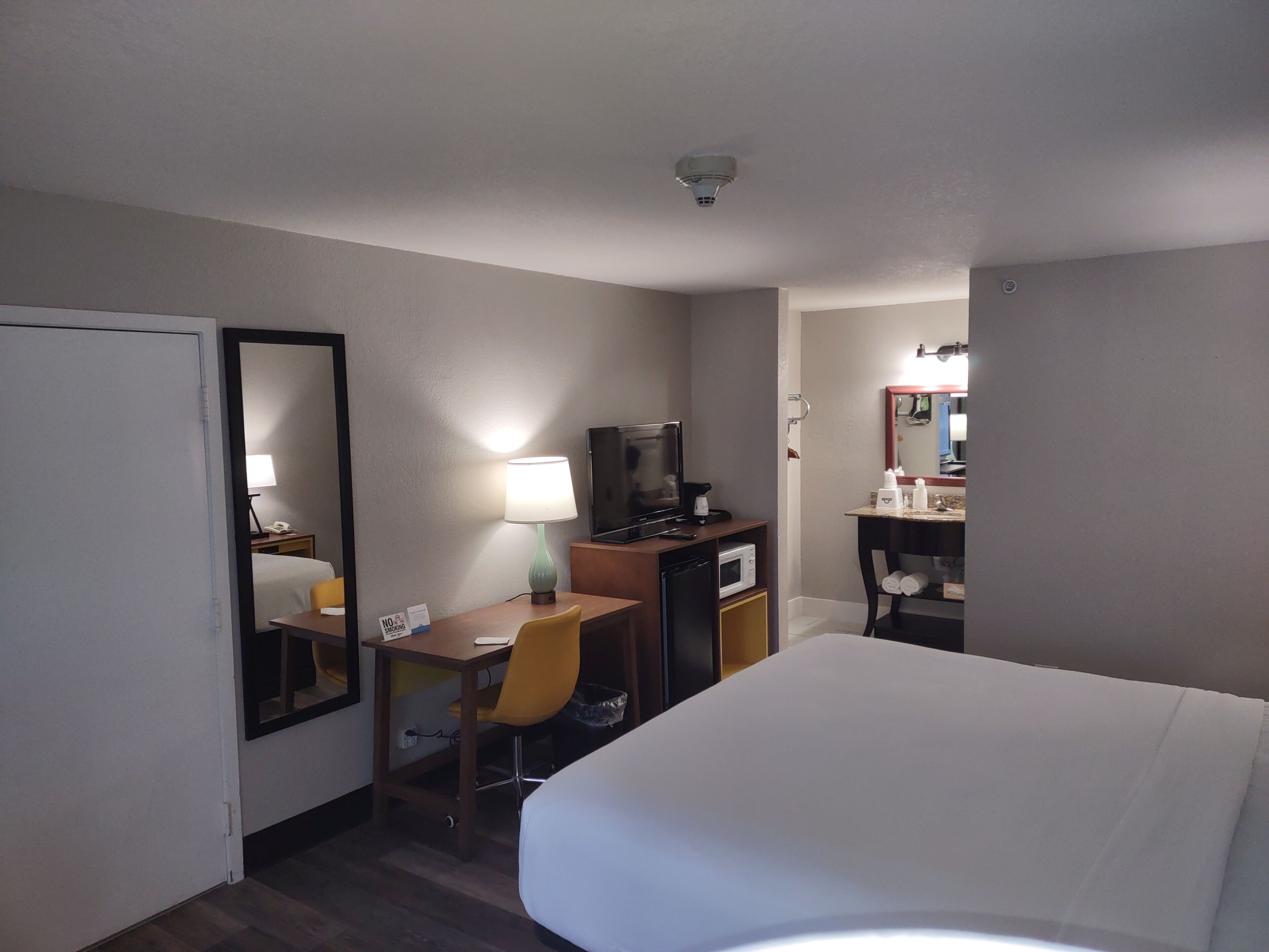 Habitación de hotel con cama, escritorio con silla, televisión, espejo, lámpara, microondas y estante pequeño con artículos de tocador.