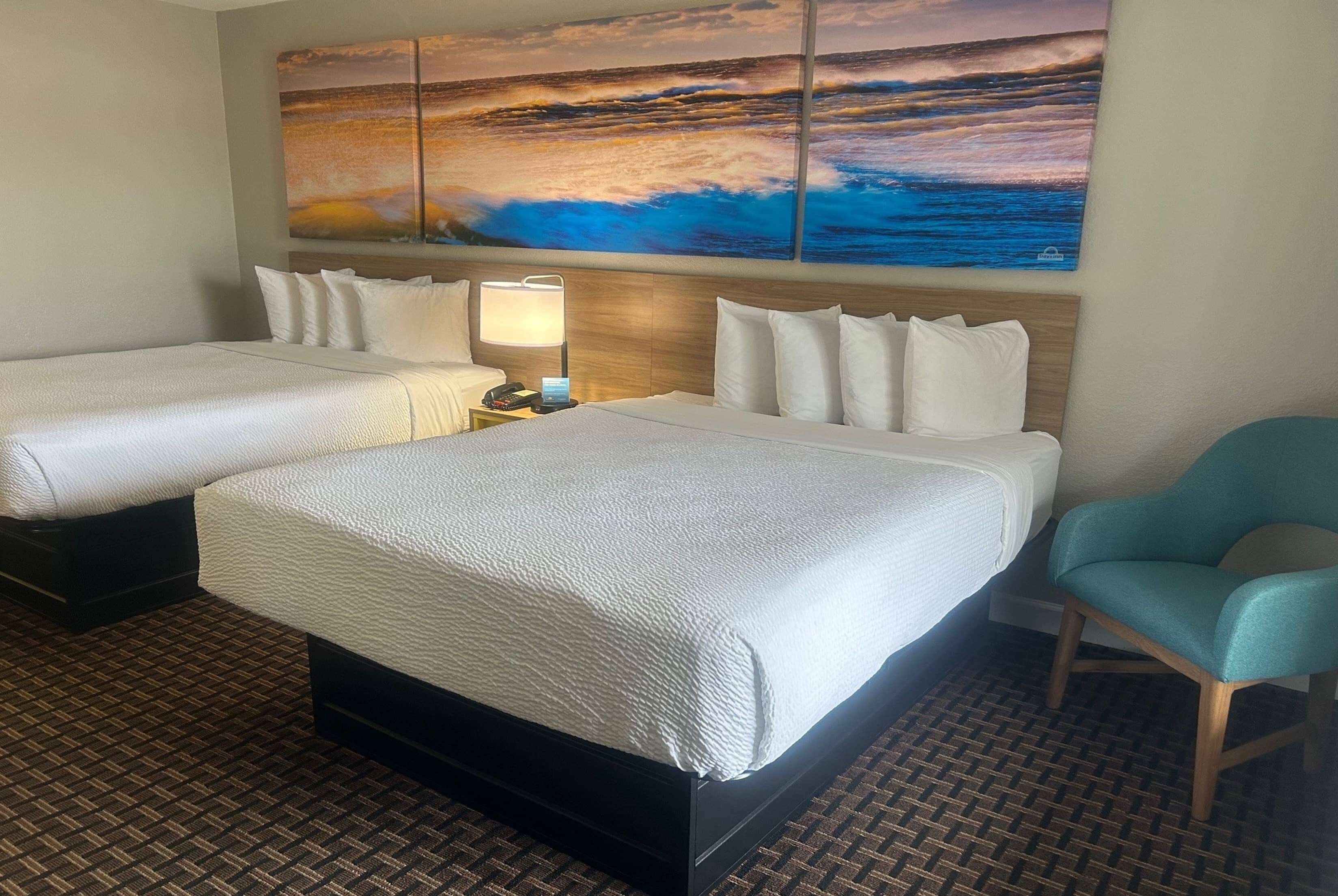 A hotel room with two beds, a nightstand with a lamp, a chair, and a large painting of a seascape on the wall.