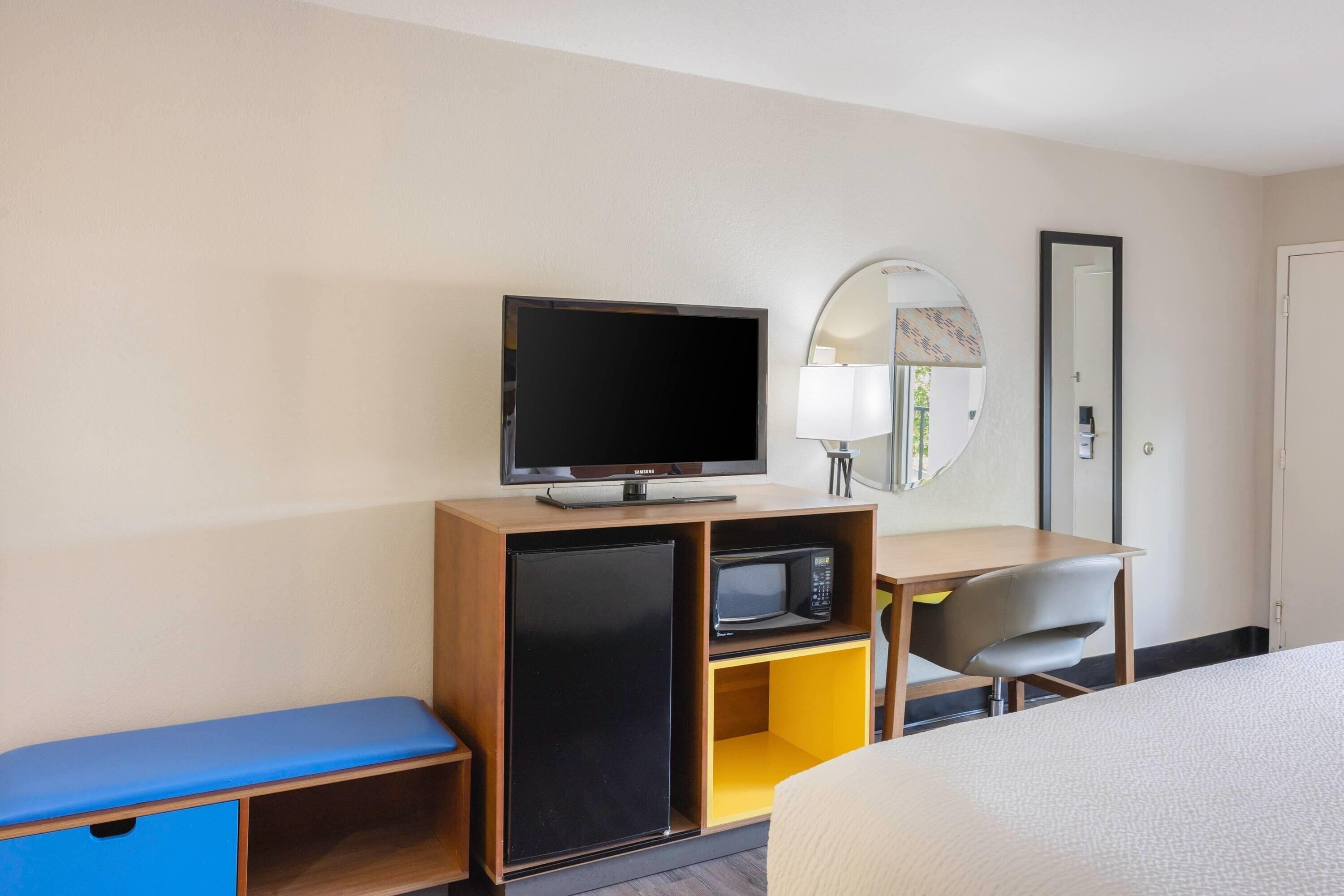 A hotel room with a bed, a desk with a television, a microwave, a small refrigerator, a chair, and a mirror.