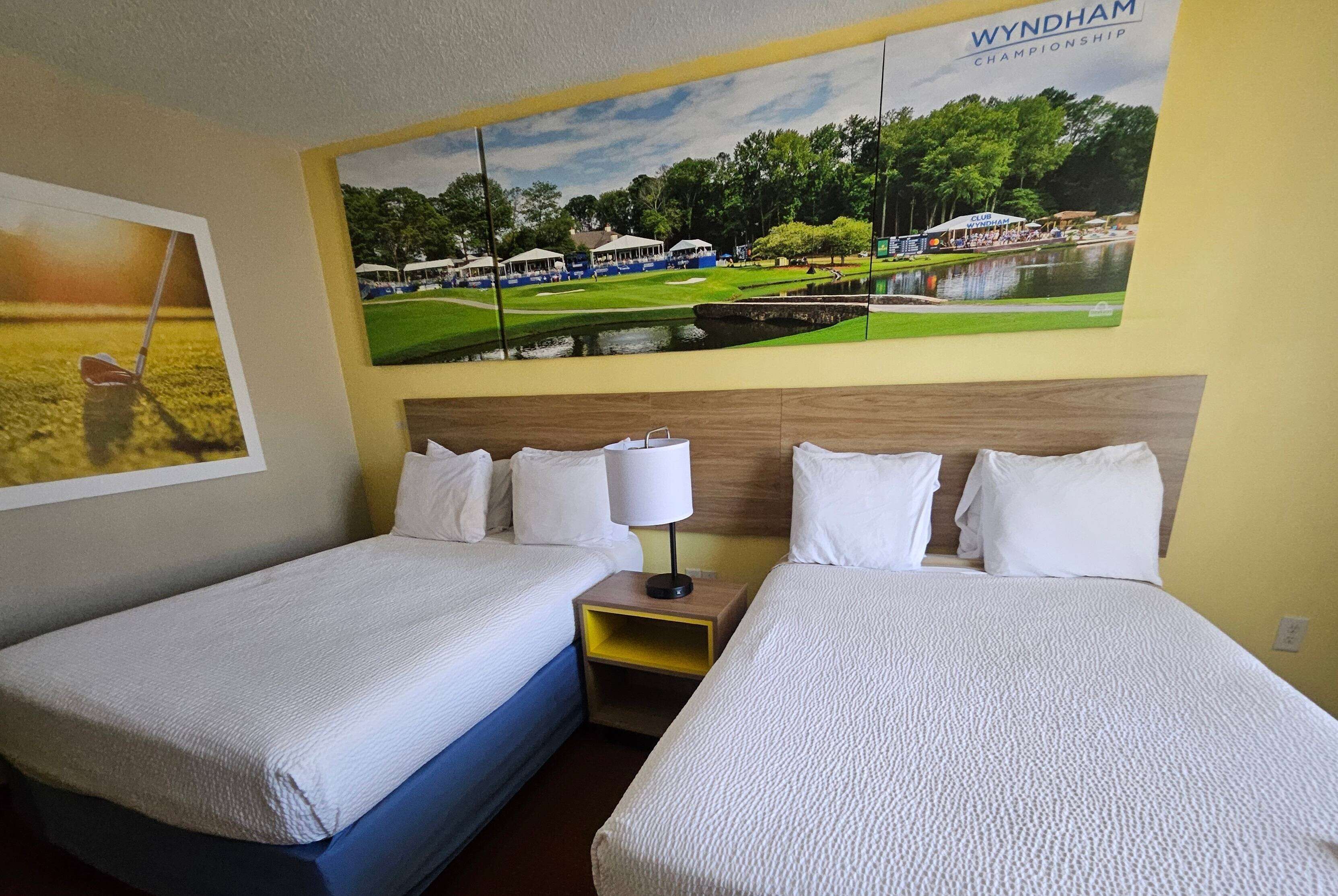 A hotel room with two beds, a nightstand with a lamp, and a large wall mural of a golf course.