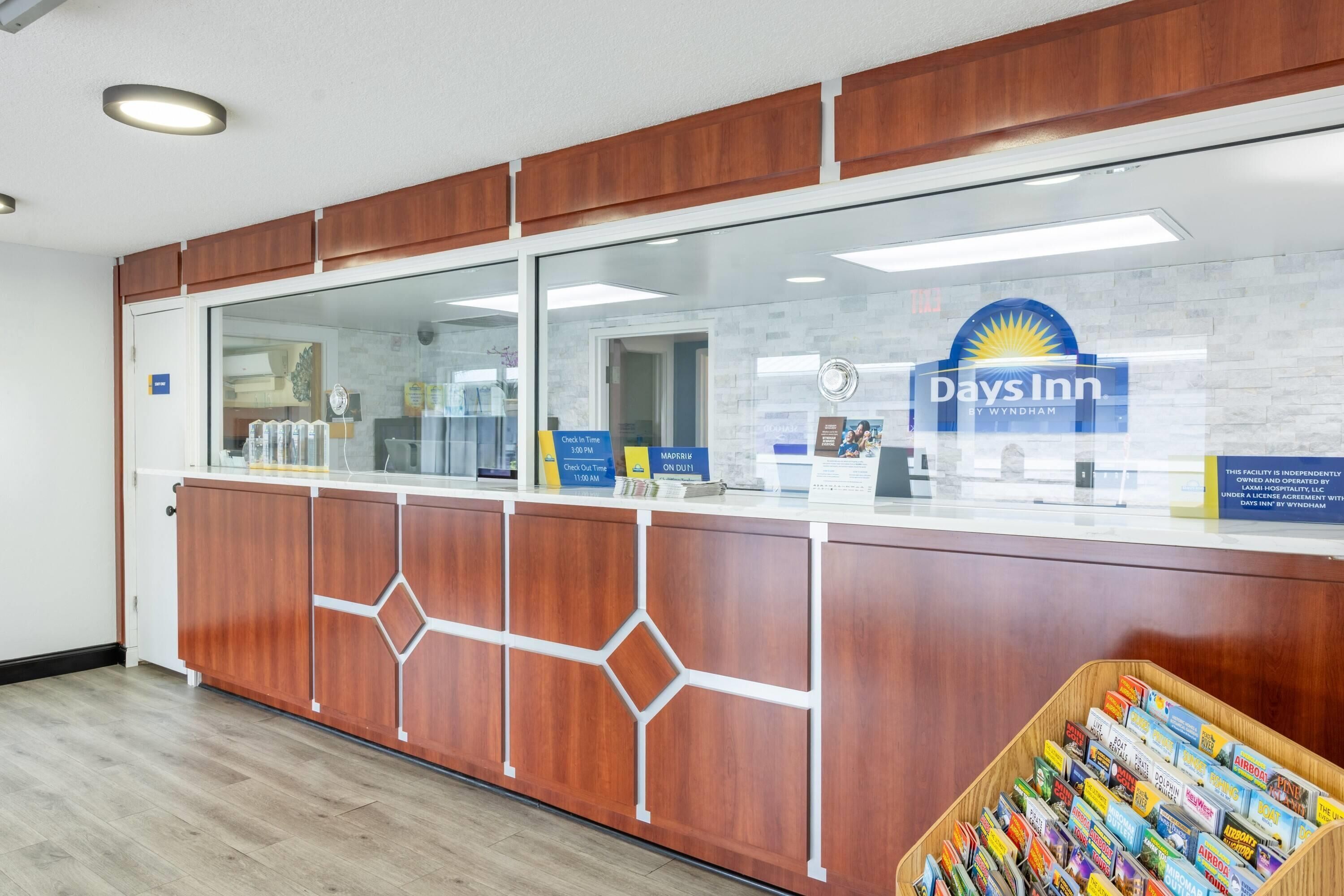 Un mostrador de recepción con separador de vidrio, el logotipo de Days Inn y un expositor para guías de viaje.