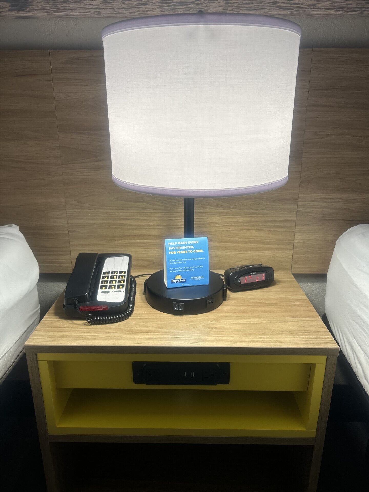 Un mesita de noche de hotel con una lámpara, un teléfono, un reloj digital y un cartel que promueve el ahorro de agua.
