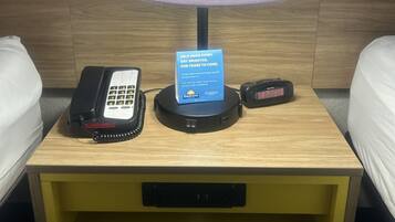 Un mesita de noche de hotel con una lámpara, un teléfono, un reloj digital y un cartel que promueve el ahorro de agua.