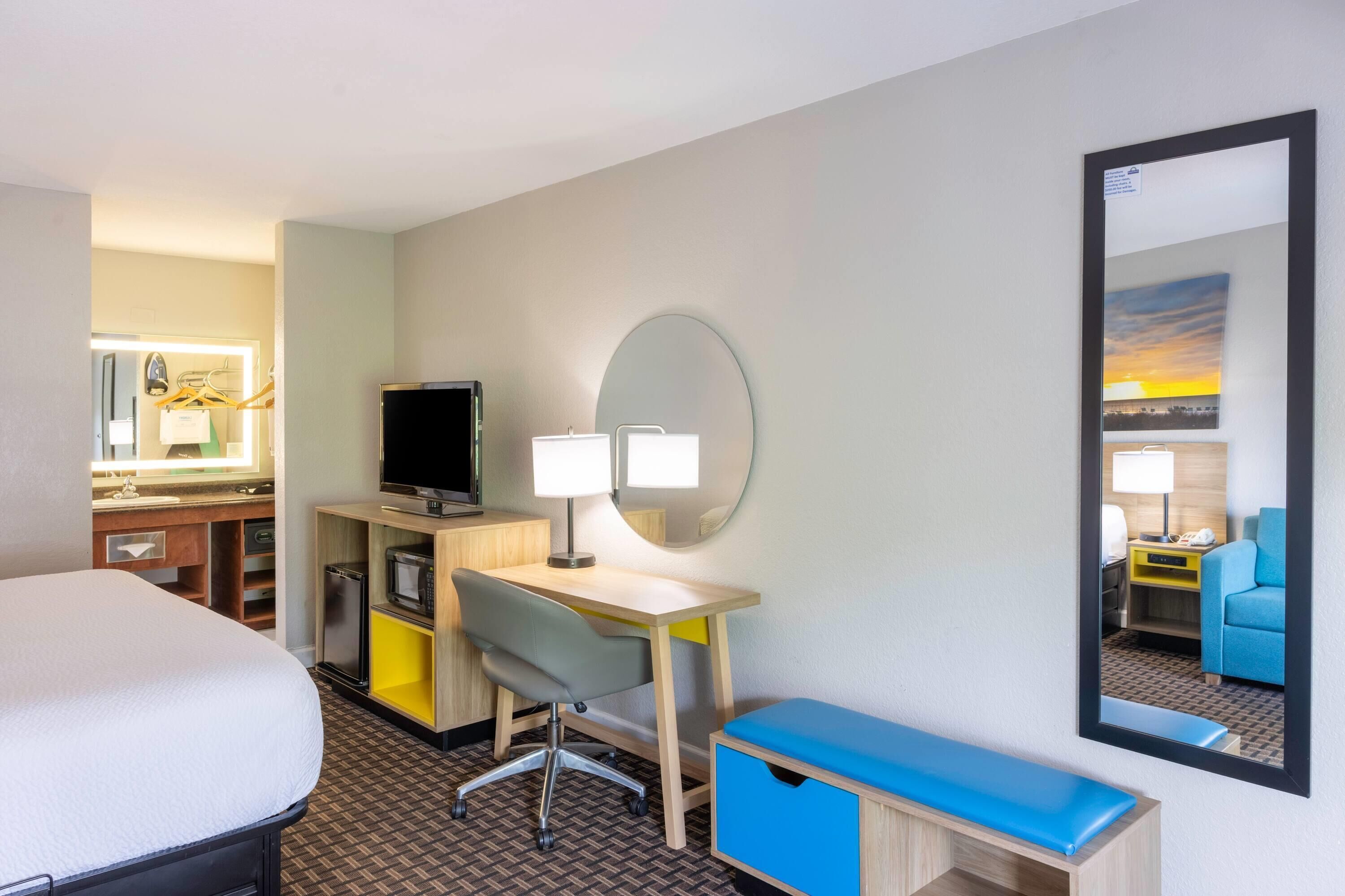 Quarto de hotel com cama, escrivaninha, cadeira, espelho e vista para outro quarto.