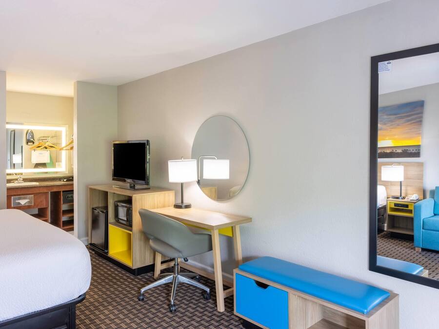 A hotel room with a bed, desk, chair, mirror, and a view of another room.